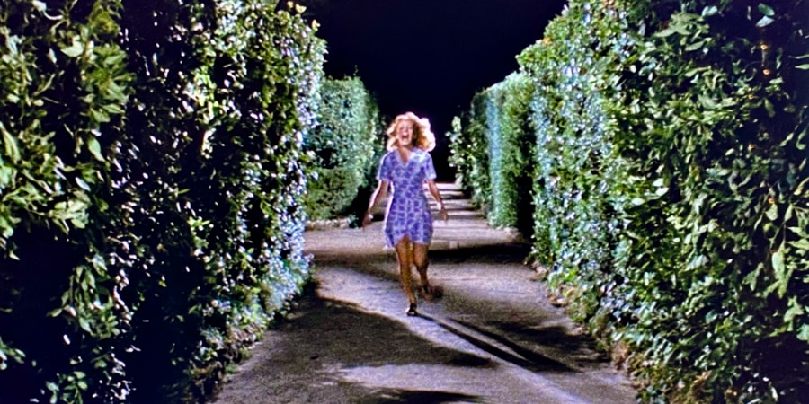 A woman runs screaming towards us with high hedges on either side.